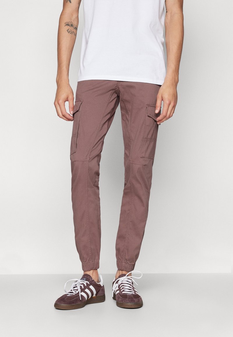 Man som bär mauve cargo joggerbyxor och lila Adidas-sneakers med vita ränder, stående mot en enkel ljus bakgrund.