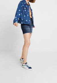 Žena oblečená v modré džínové bundě s puntíky, oranžovém topu, tmavě džínové minisukni a modro-bílých teniskách, stojící s jednou nohou vpřed.