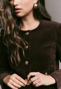 Brown corduroy jacket with a round neckline, featuring gold ornate buttons and flared sleeves. Long wavy hair partially visible.