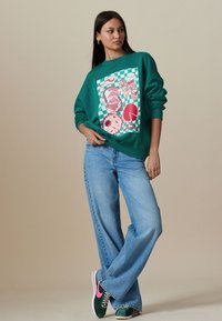 Zelená mikina s grafickou potlačou potravín na kockovanom pozadí, doplnená modrými džínsami s širokými nohavicami a zelenými teniskami s ružovými akcentmi.