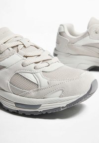 Beige Laufschuhe aus einer Kombination von Mesh- und Wildledermaterialien, mit strukturierten Schnürsenkeln und einer konturierten Gummisohle. Akzentuiert mit weißen Details.