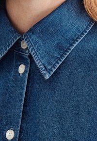Camicia di denim con colletto classico, colore azzurro chiaro e tessuto texturizzato. Presenta bottoni bianchi e dettagli di cucitura visibili.