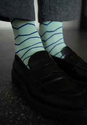 Person trägt hellgrüne Socken mit blauen Zickzackstreifen und schwarze Lederloafer auf dunklem Fliesenboden.