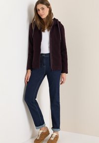 Veste en polaire bourgogne avec capuche, fermeture éclair noire et cordon de serrage ; associée à un jean en denim foncé et des baskets marron clair.
