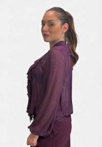 Blusa viola trasparente con maniche lunghe, dettagli di volant sul davanti e colletto. Abbinata a pantaloni viola coordinati.