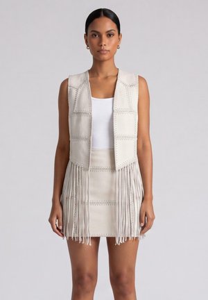 Donna che indossa un gilet beige senza maniche e una gonna abbinata con dettagli in frange sopra una canotta bianca, in piedi davanti a uno sfondo semplice.