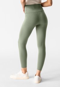 Leggings a vita alta, aderenti, di colore verde oliva chiaro, realizzati in un tessuto liscio e elastico; abbinati a scarpe sportive bianche.