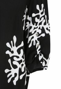 Schwarze Stoffbluse mit einem weißen korallenähnlichen Muster; verfügt über Puffärmel mit Knopfdetail und glatter Textur.
