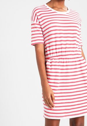 Frau trägt ein kurzärmeliges, weißes Kleid mit horizontalen roten Streifen und elastischem Taillenband vor einem einfarbigen weißen Hintergrund.