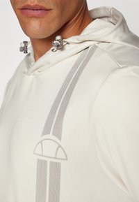 Gai bēšs kapucis ar faktūra apdari, ar pelēku abstractu rakstu un regulējamu pelēku auklu aparatūru kapuci.