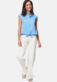 Blusa azzurra chiara a maniche corte con colletto, orlo cinched, abbinate a pantaloni bianchi a gamba larga e sandali con fasce, su uno sfondo neutro.
