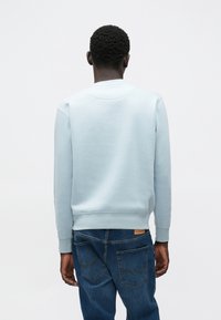 Gaiši zila džempera ar gludu tekstūru, ribveida aprocēm un apaļu kakla izgriezumu, komplektā ar zilām džinsu biksēm ar aizmugurējām kabatām.