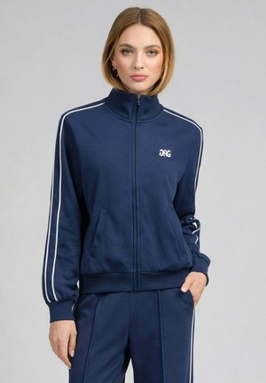 Jeune femme portant une veste de survêtement zippée bleu marine et un pantalon assorti avec des bandes blanches sur les manches et les côtés, debout devant un fond uni.