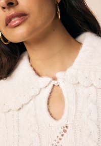 Beli pleteni pulover z robom v obliki valov, odprtino v obliki ključavnice, pritrjeno z bisernim gumbom, in teksturiranimi vzorci pletenice.