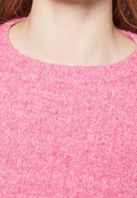 Rosa stickad tröja med ribbad textur och rund halsringning. Tyget ser mjukt och något luddigt ut med en rik färgdjup.