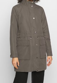 Grå knälång jacka i syntetmaterial med hög krage, tryckknappar och två framfickor. Har justerbar dragsko i midjan.