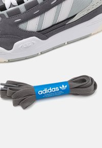 Szary sneaker marki Adidas z cholewką z zamszu i siateczki, z białymi trzema paskami i gumową podeszwą. Zawiera dopasowane szare sznurowadło z niebieską metką.