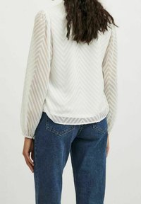 Valkoinen paita, jossa on teksturoidut, läpikuultavat zigzag-hihat. Takana on pyöreä helma, ja malli on yhdistänyt sen sinisiin farkkuihin.