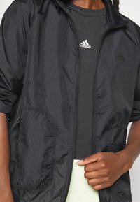 Chaqueta negra con cremallera y patrón texturizado, con bolsillos laterales y capucha ajustable, llevada sobre una camiseta negra acanalada.