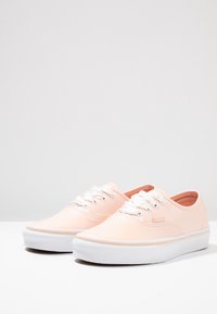 Jasnoróżowe trampki z płótna z białymi sznurowadłami, o niskim kroju i teksturowanej gumowej podeszwie. Charakterystyczne logo z boku.