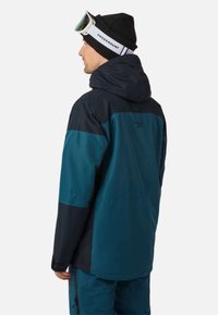 Man wearing blue and black ski jacket, matching pants, black beanie, and white Swedemount ski goggles, facing left on a white background.