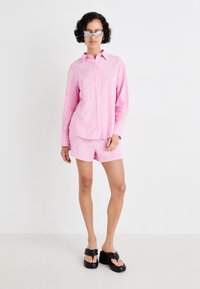 Camisa abotoada rosa às riscas combinada com calções iguais. Modelo usa sandálias de plataforma pretas e óculos de sol. Design simples e Casual.