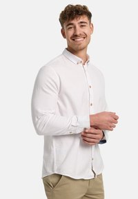 Weißes langärmliges Hemd mit Knöpfen aus leichtem Stoff, braunen Knöpfen und abgerundetem Saum. Kombiniert mit hellbeigen Hosen.