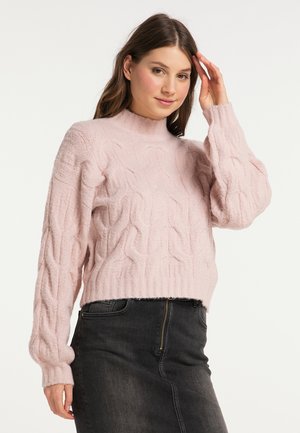 Frau berührt ihr Haar, trägt einen hellrosa Zopfmuster-Pullover und einen hoch taillierten dunkelgrauen Rock mit Reißverschluss vorne.