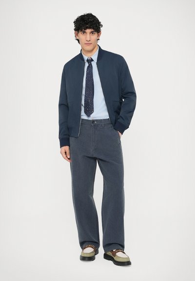 Námornícka bomber bunda cez svetlomodrú košeľu, v kombinácii so sivými širokými kordovými nohavicami a hnedo-krémovými topánkami, s vzorovanou kravatou.