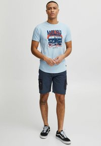 Un tricou bleu deschis cu un grafic de autobuz vintage și textul "Malibu", asortat cu pantaloni scurți negri cu buzunare laterale și adidași negri.
