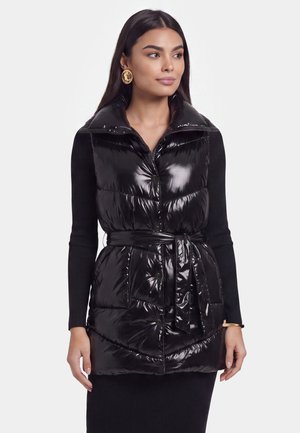 Frau trägt eine schwarze glänzende Steppweste mit Gürtel über einem schwarzen Langarmshirt, ergänzt durch goldene Ohrringe und ein Armband.