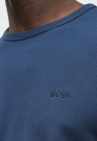 Camisola de gola redonda azul texturada com um discreto logótipo "BOSS" em relevo no peito. O material é macio e respirável.