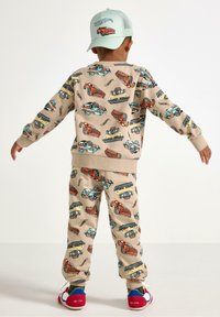 Beige bomulds sweatshirtsæt med bukser, der har et flerfarvet bilprint. Udsmykket med ribbede manchetter og en lyseblå kasket med logo.