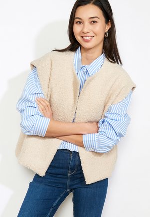 Junge Frau mit glattem dunklem Haar, trägt silberne Creolen, eine beige ärmellose strukturierte Weste über einem blau-weiß gestreiften Hemd und blaue Jeans.