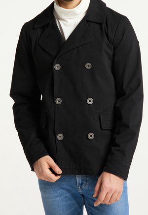 Homme portant une veste noire croisée à six boutons avec poches à rabat sur un col roulé blanc et un jean bleu.