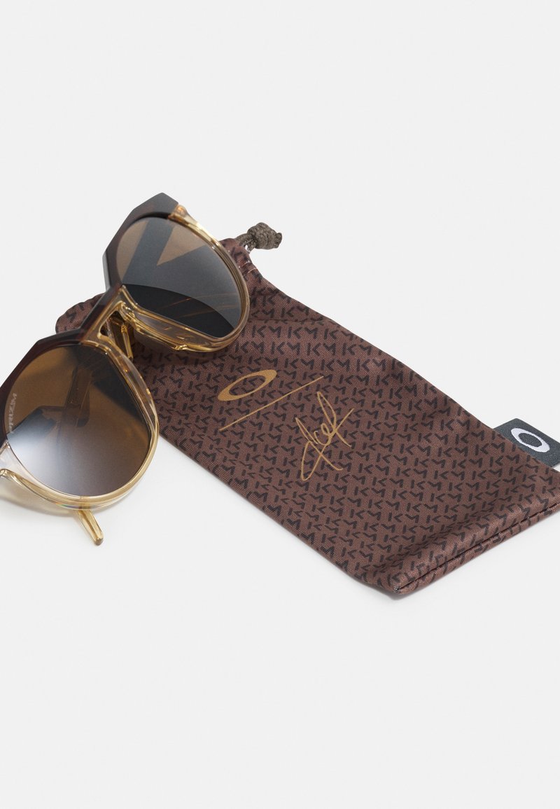 Lunettes de soleil marron avec des montures angulaires, verres marron et accents dorés, posées sur une pochette douce marron à motifs avec un logo doré.