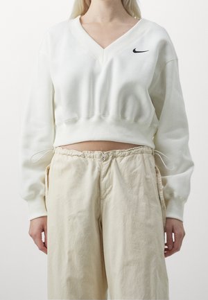 Kobieta ubrana w białą skróconą bluzę z dekoltem w serek i logo Nike oraz beżowe, luźne spodnie z elastycznym pasem.