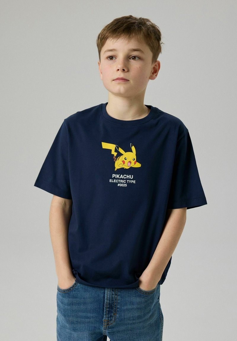 Αγόρι με μπλε ναυτικό T-shirt με γραφικό Pikachu και μπλε τζιν, στέκεται με τα χέρια στις τσέπες μπροστά από απλό φόντο.