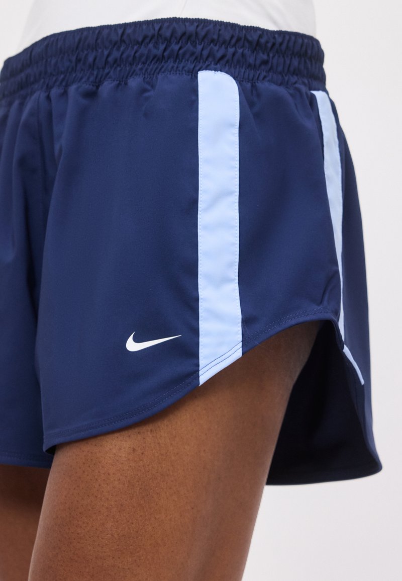 Primer plano de una persona que lleva pantalones cortos deportivos azul marino con rayas verticales azul claro y un logo blanco de Nike en el muslo izquierdo.