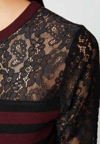 Zwarte kant top met bloempatronen, bordeauxrode en zwarte gestreepte onderkant, ribgebreide kraag en doorschijnende mouwen.