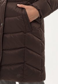 Bruine pufferjack met een geborduurd ontwerp, voorzien van zijzakken en contrasterende afwerking. Logo detail nabij de onderkant aan de linkerkant.