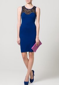 Tajtat blå klänning med en svart spetsmönstrad halsringning. Modellen håller en lila kuvertväska och bär blå klackar. Slät tyg, knälång design.