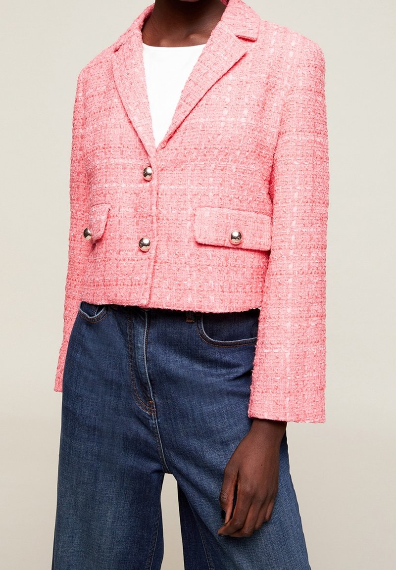 Różowy blezer z tweedu z klapami, dwoma guzikami i przednimi kieszeniami. Długie rękawy i krótki fason, zestawiony z niebieskimi spodniami dżinsowymi.