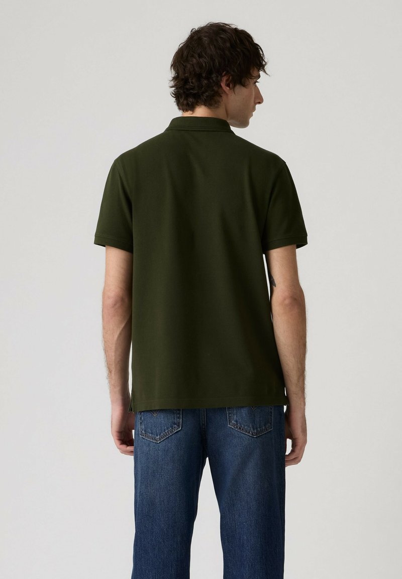 Homme portant un polo vert foncé à manches courtes et un jean bleu, vu de dos sur un fond clair uni.