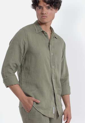 Giovane uomo che indossa una camicia di lino verde oliva con bottoni e maniche arrotolate, in piedi con una mano in tasca, guardando leggermente di lato.