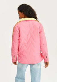 Růžová prošívaná bunda se žlutým límcem a zlatými akcentními pruhy; má zipové manžety a vzor ve tvaru šípu. Zobrazená zadní část.