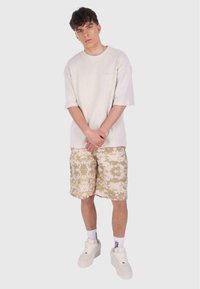 Lys grå oversized struktureret t-shirt og beige camo shorts, med hvide sneakers. Bemærkelsesværdige detaljer inkluderer ribbet stof og logo accenter.