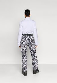 Cămașă albă cu mânecă lungă, asortată cu pantaloni cu imprimeu leopard albastru și bej. Curea neagră cu detaliu logo. Modelul stă cu spatele la privitor.