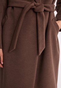 Brązowy płaszcz z tkaniny o fakturze, wyposażony w zawiązywany pasek, kieszenie po bokach oraz prosty design bez widocznych zapięć.
