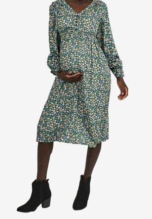 Robe de maternité florale à manches longues, fond vert, motif floral multicolore, longueur genou, associée à des bottines noires.